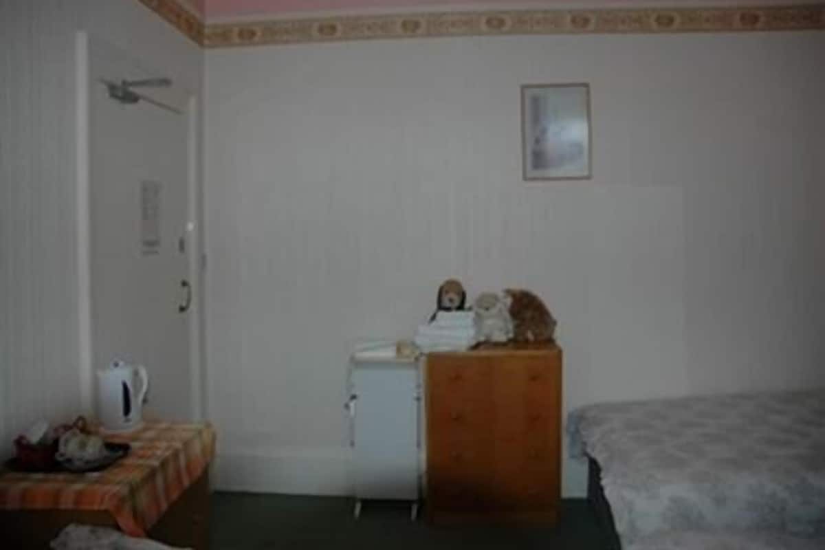A room with a bed, a small table with a kettle, a cabinet, and a framed picture on the wall.