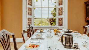 A dining room with a large window, a table set for breakfast, and a view of a green field. - Smeaton Farm (Saltash)
