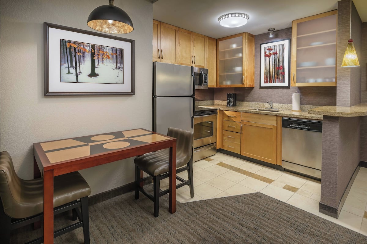 A compact kitchen with wooden cabinets, stainless steel appliances, a dining table with chairs, and a framed picture on the wall.