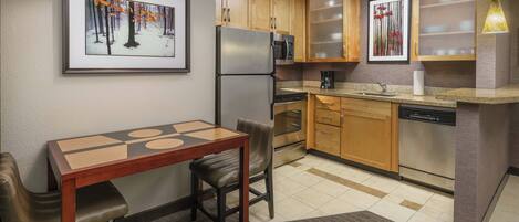 A compact kitchen with wooden cabinets, stainless steel appliances, a dining table with chairs, and a framed picture on the wall.