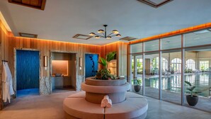 Un área de spa con un espacio de descanso circular, un mostrador de recepción y un ventanal grande con vistas a una piscina interior.