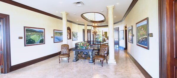 A spacious hallway with a marble floor, framed landscape paintings, and a central decorative table with chairs.