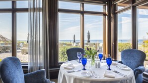 Una mesa de comedor con sillas azules, dispuesta para dos, con vistas a un paisaje marino y un patio con mobiliario exterior.