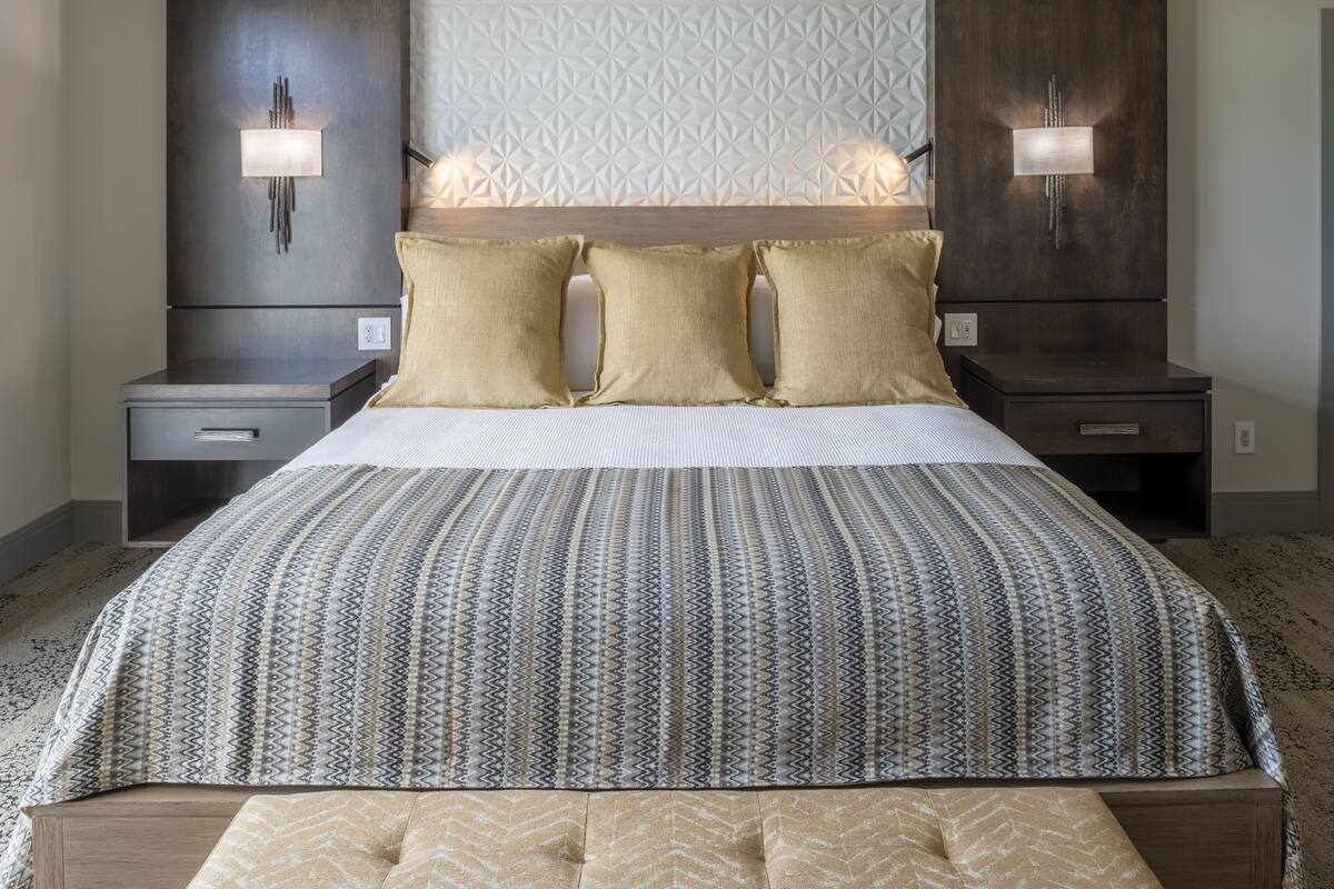 Una cama bien hecha con una colcha estampada y tres almohadas, flanqueada por mesitas de noche con lámparas de pared.