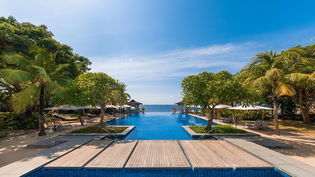 A clear view of a swimming pool with a wooden deck, surrounded by palm trees and lounge chairs. - Crimson Resort and Spa Mactan (Lapu-Lapu)