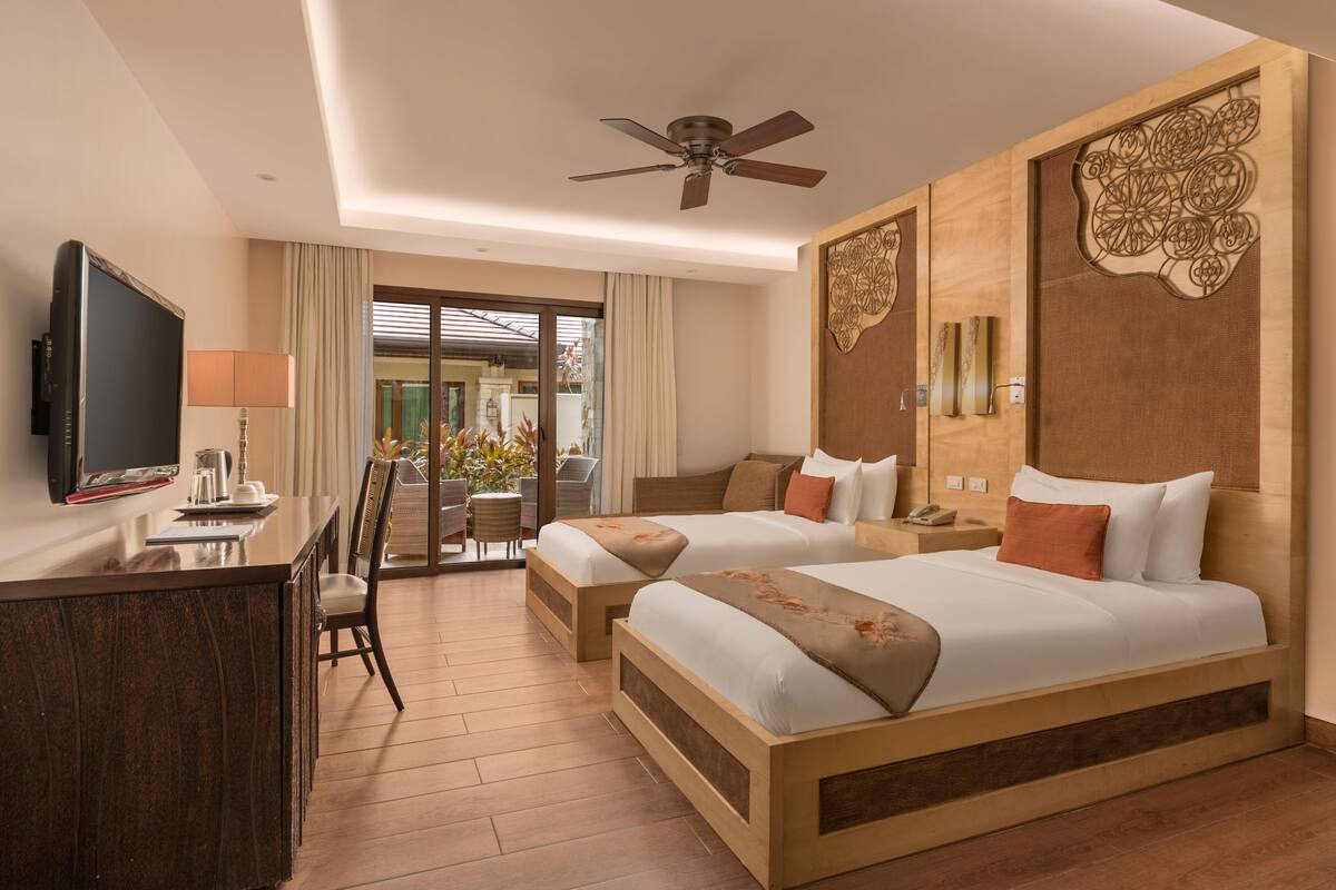 Une chambre d’hôtel avec deux lits, un bureau avec une télévision, un ventilateur de plafond et une vue sur un jardin.