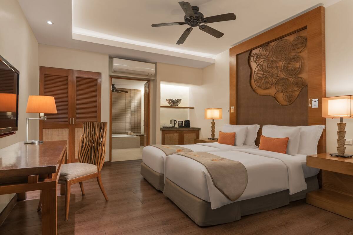 Une chambre d’hôtel comprenant un lit, deux tables de chevet, un bureau, une chaise, un ventilateur de plafond et un élément décoratif mural.