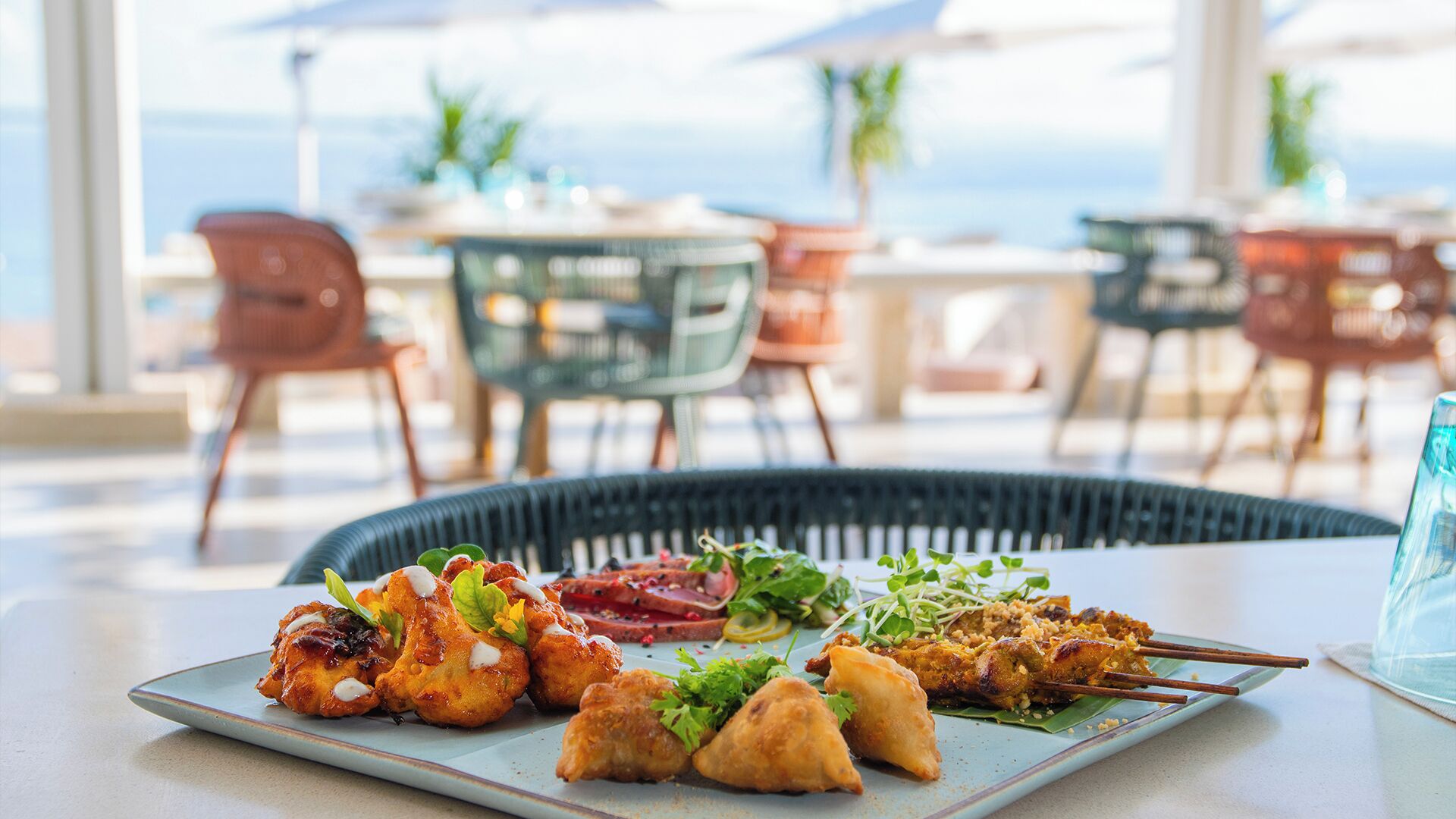 Una mesa de comedor con diversos platos, incluyendo alimentos fritos y brochetas, en una terraza con sillas y mesas al fondo.