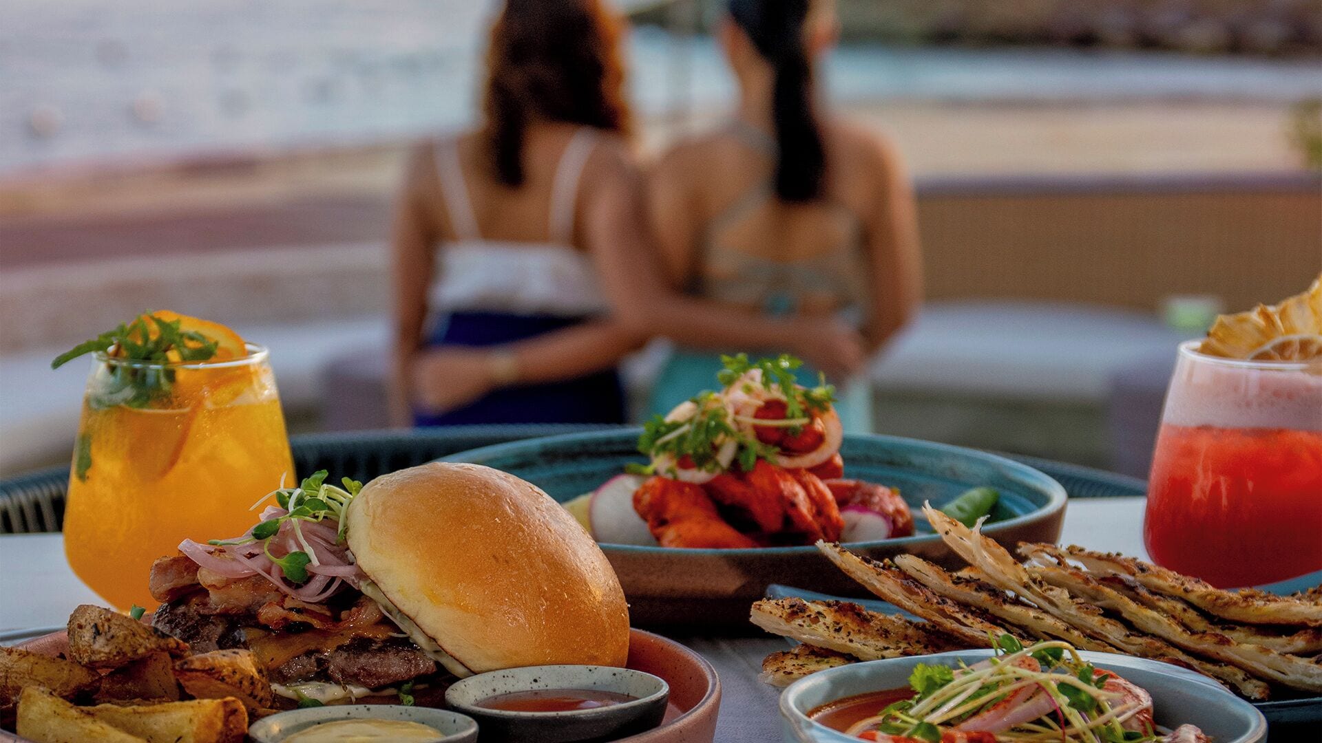 A dining table with a variety of dishes including a burger, grilled chicken, and seafood, along with drinks and condiments.