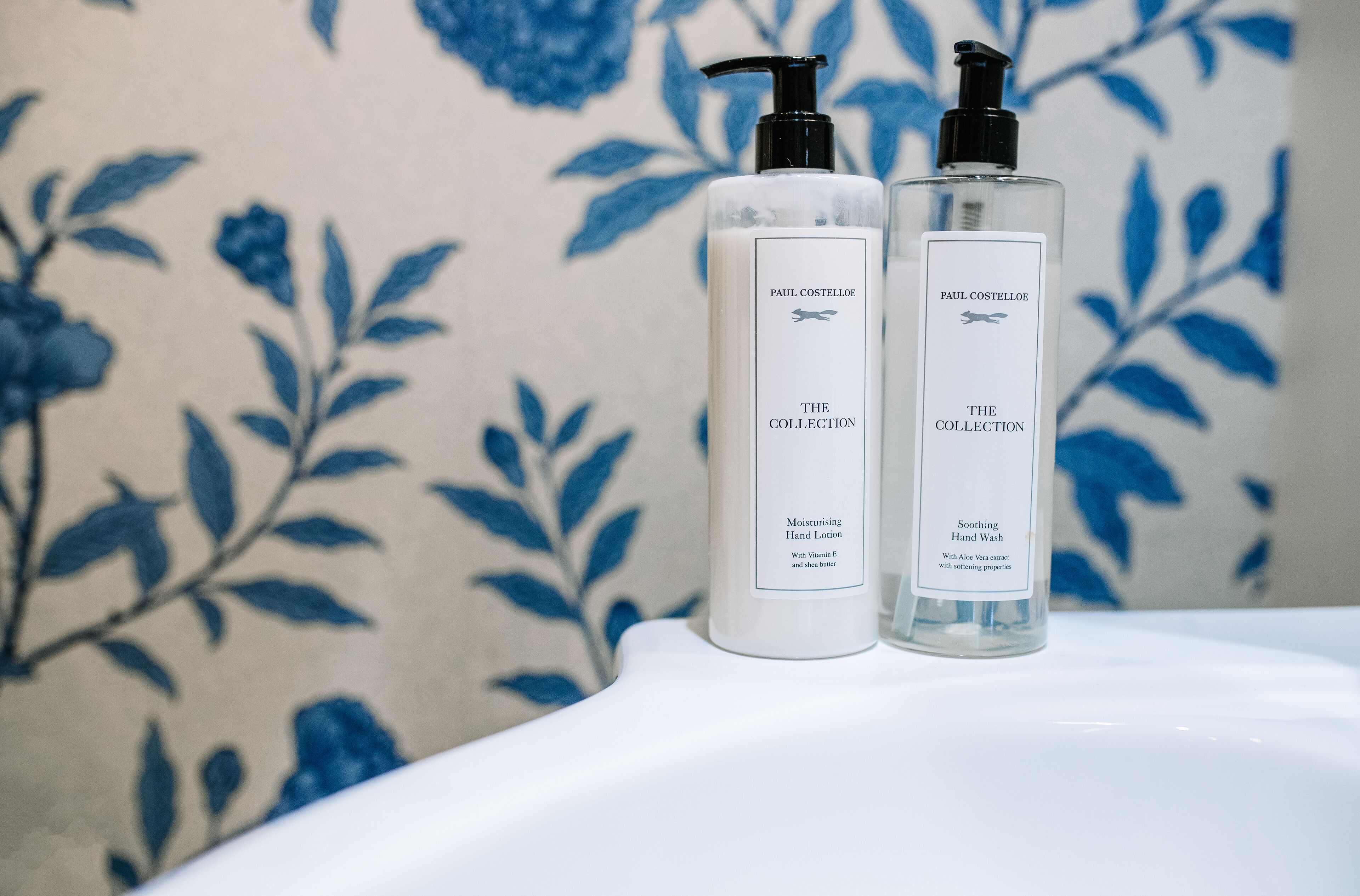 Two Paul Costello The Collection hand wash bottles on a white surface with a floral patterned background.