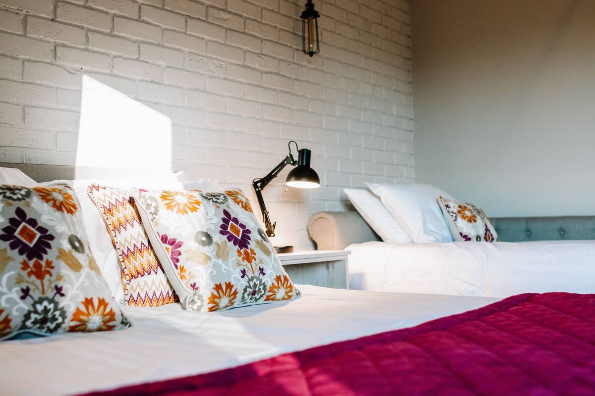 A hotel room with a white brick wall, a bed with a colorful floral pillow, a bedside table with a lamp, and a nightstand with a lamp.