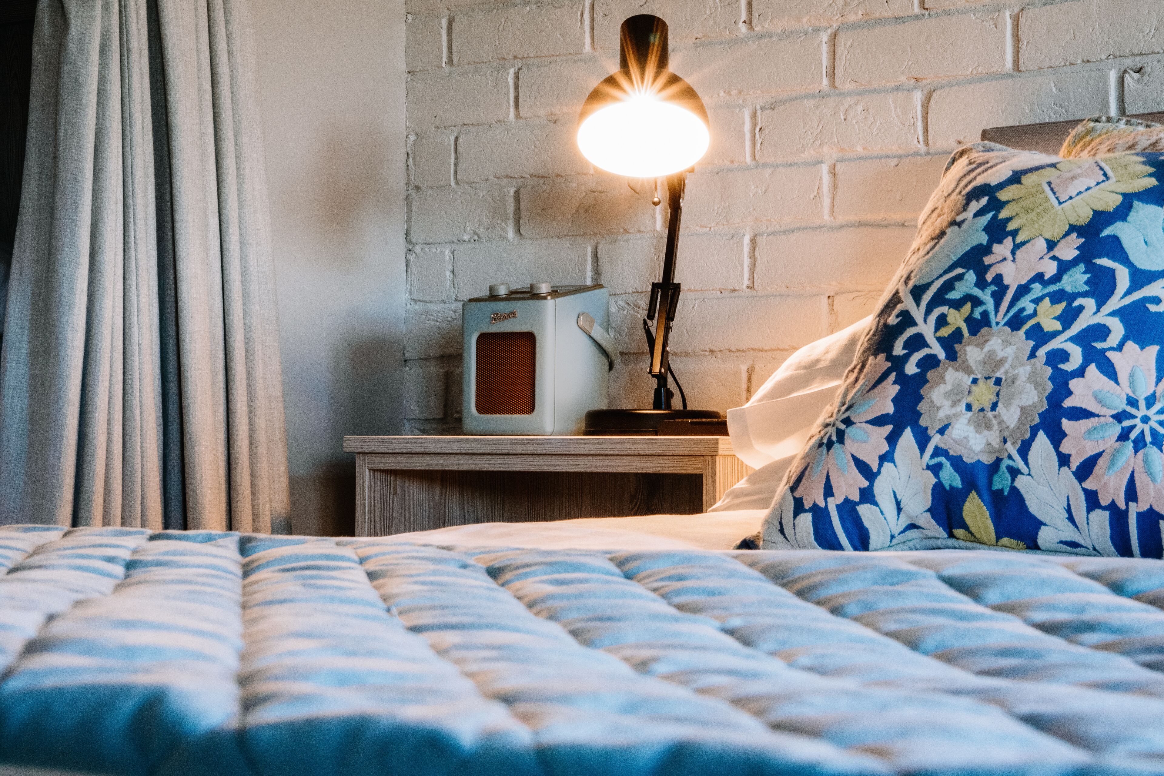 Una cama con una colcha de diseño azul y blanco, un almohadón blanco con motivos florales, una lámpara de noche y una radio antigua sobre una mesita de noche de madera, todo contra un muro de ladrillo.