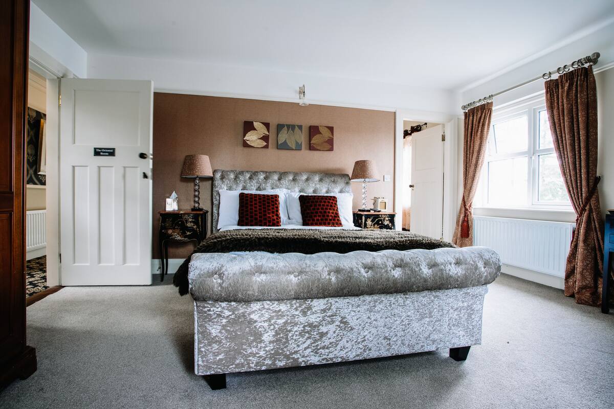 A bedroom with a large bed, two bedside tables, a nightstand, a bench, a window with curtains, and a door.