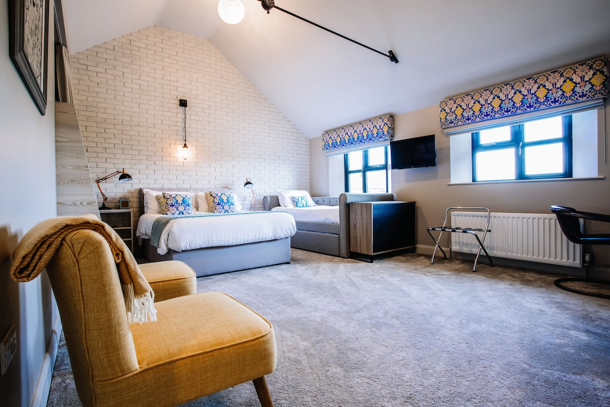 A bedroom with a sloped ceiling, a bed, a chair, a desk, a radiator, and a window with patterned curtains.