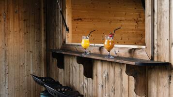 A rustic wooden cabin with a wooden bench, two metal lanterns, and a shelf with two glasses of orange juice.