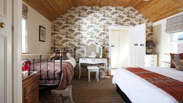 Un dormitorio con techo de madera, una cama con colcha roja y blanca, un aparador, una tocador con espejo y papel tapiz estampado.
