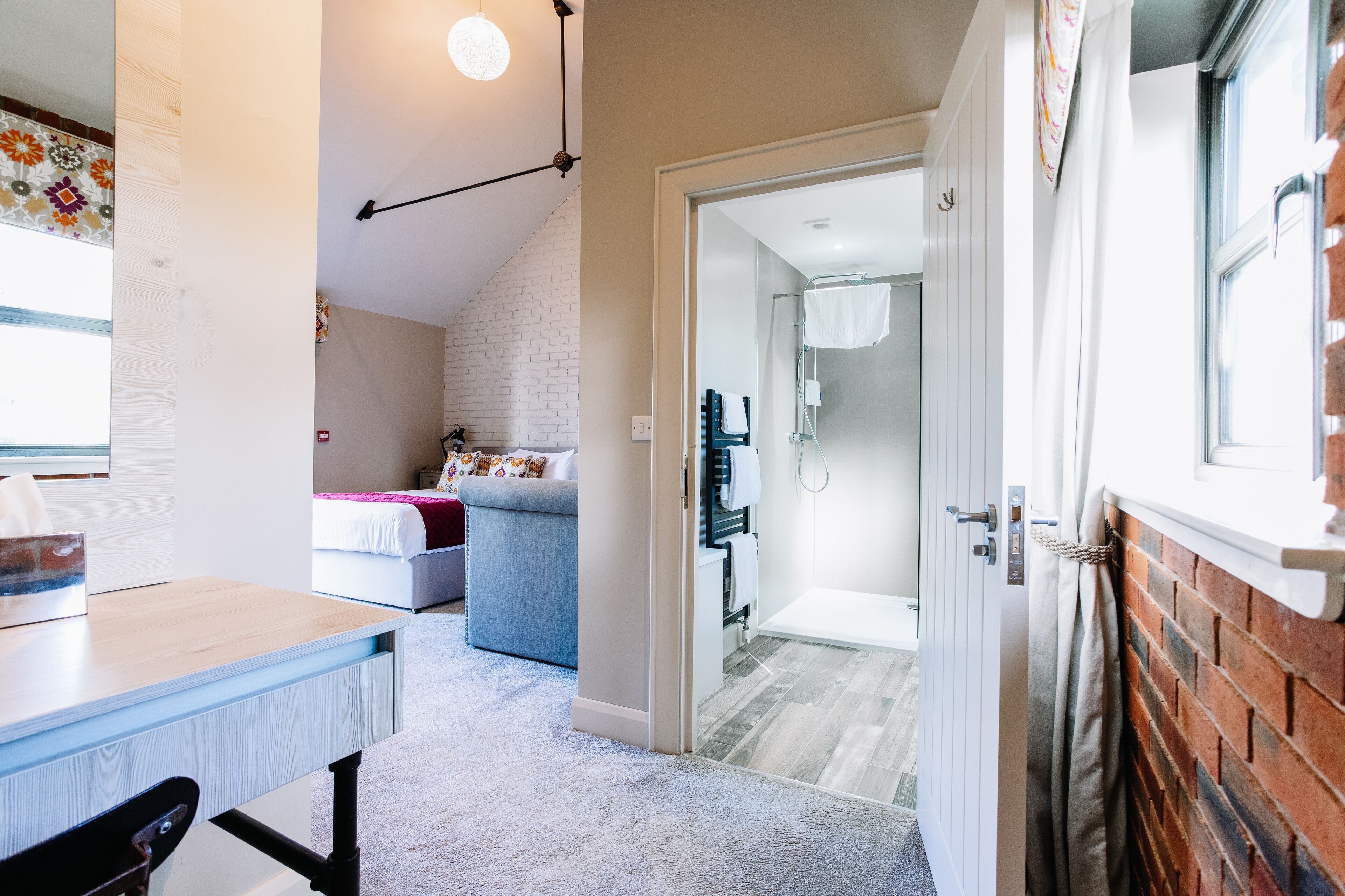Una habitación moderna con un escritorio de madera, un sofá gris y una cama. Se ve un baño con ducha y retrete a través de una puerta abierta.