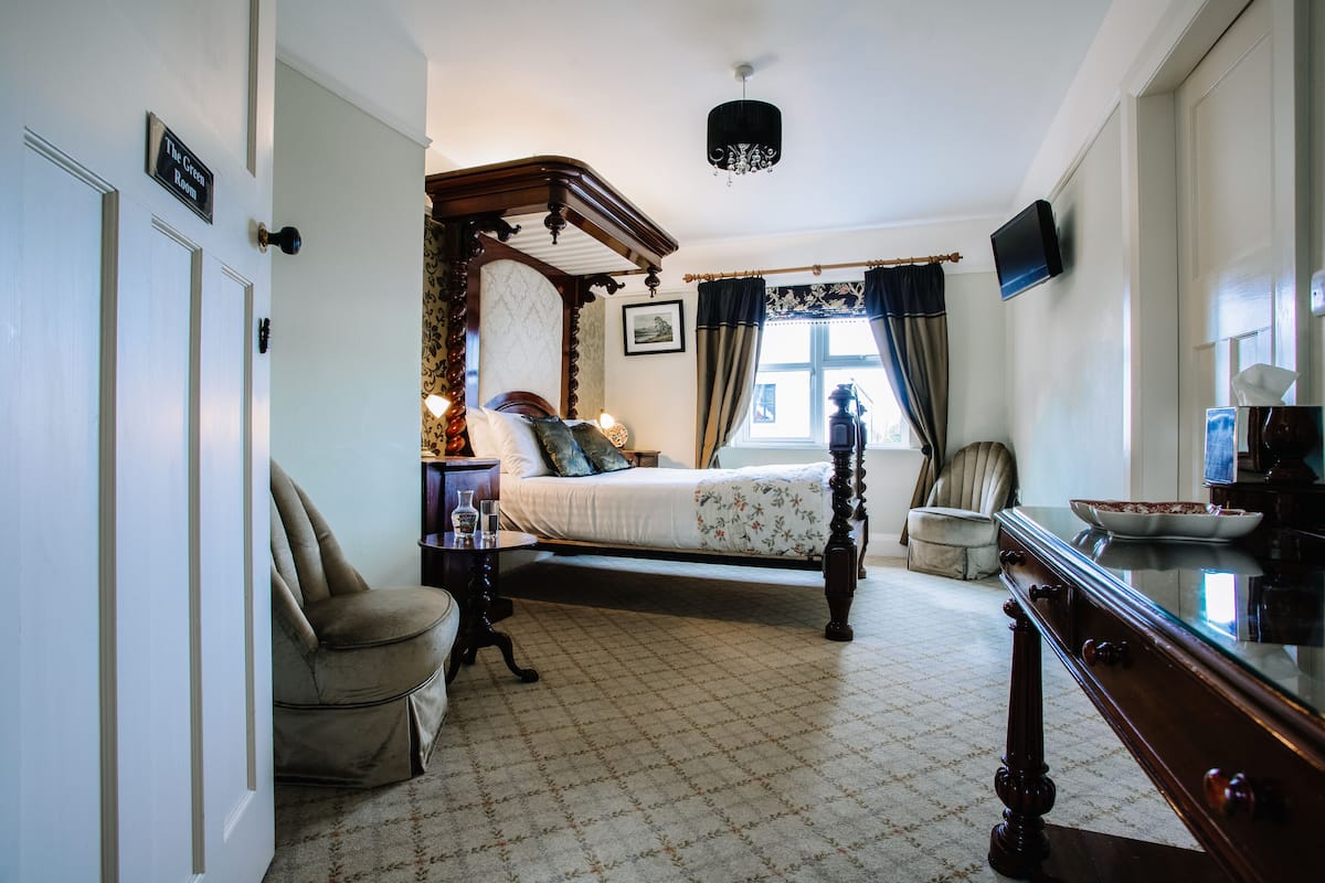 A traditional bedroom with a four-poster bed, a wooden dresser, a chair, and a television on the wall.