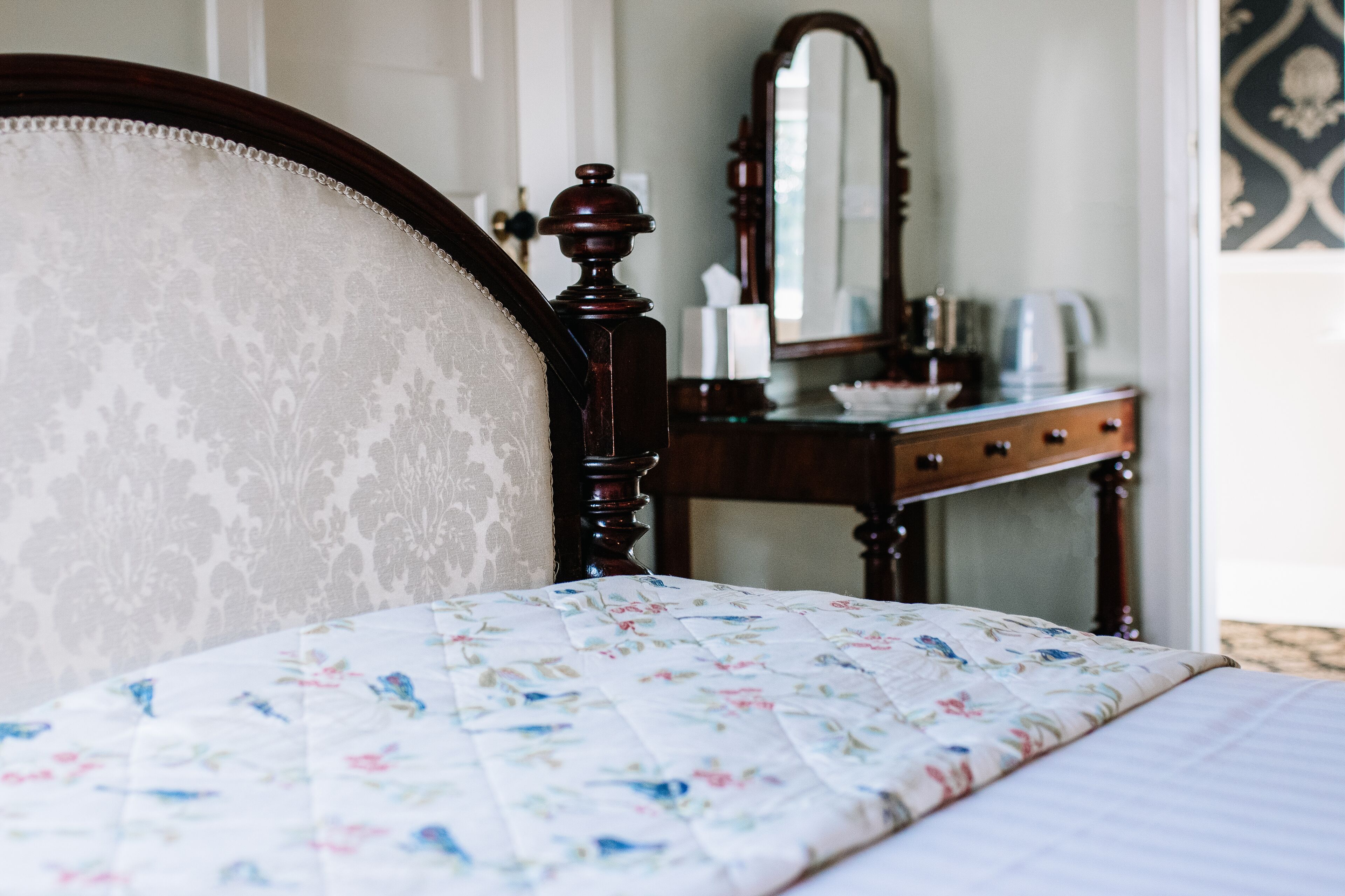 Una cama con colcha floral, cabecera de madera, cómoda con espejo y un panel decorativo en la pared.