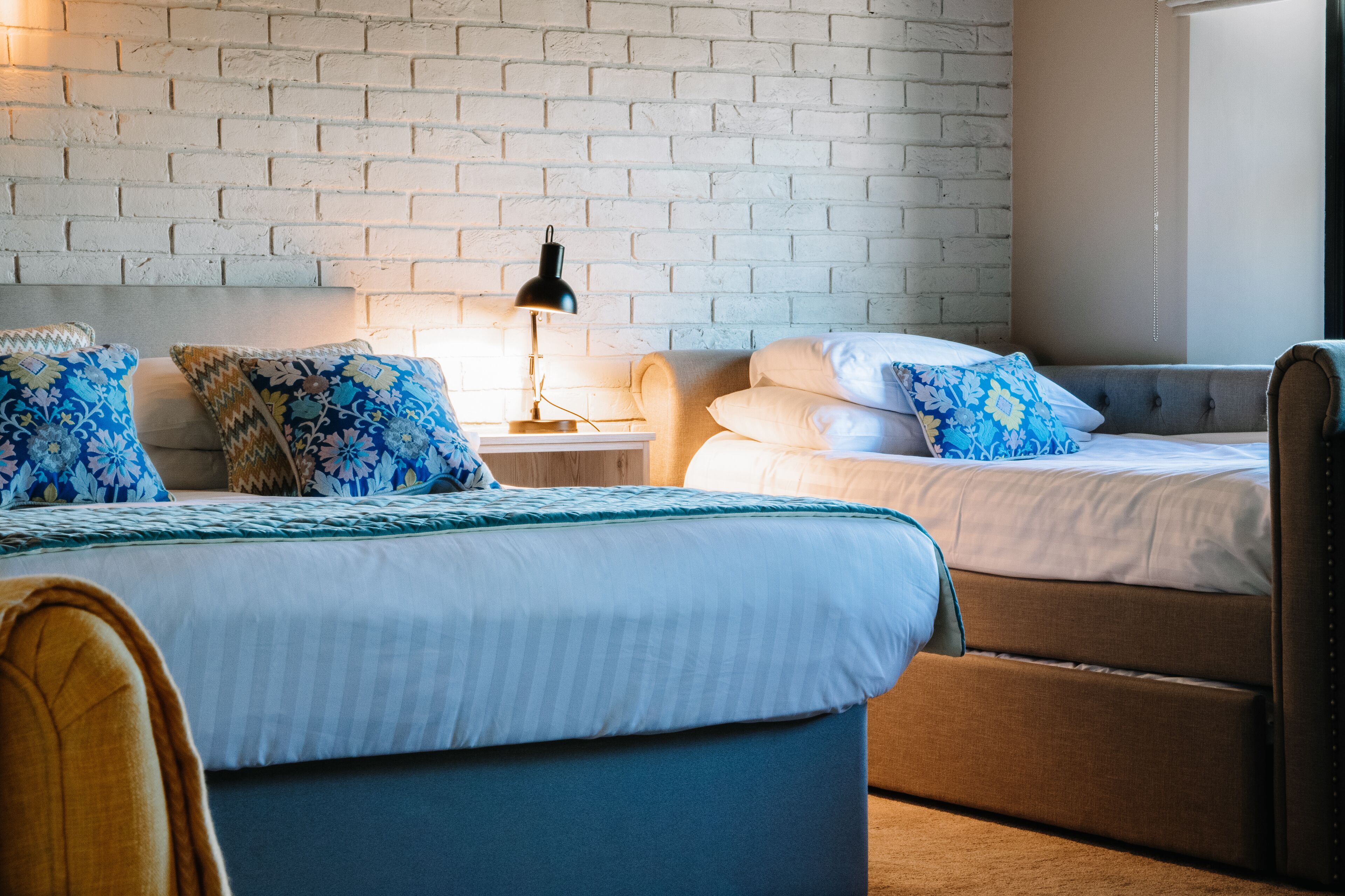 A hotel room with a brick wall, two beds with patterned bedding, a nightstand with a lamp, and a chair.
