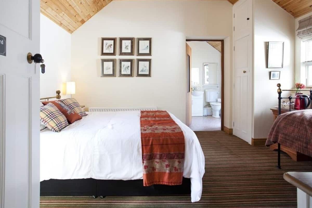 A hotel room with a bed, a wooden ceiling, framed pictures on the wall, and a bathroom visible through an open door.