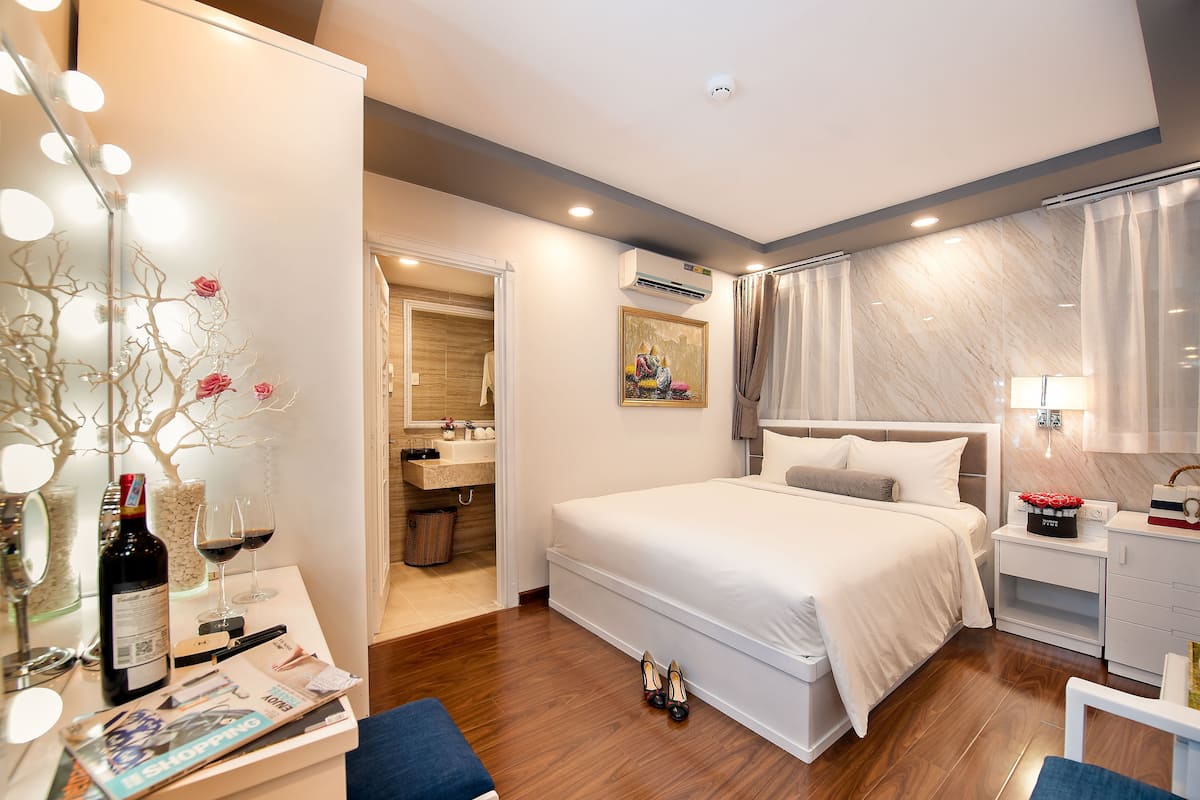 Une chambre d’hôtel moderne avec un grand lit, une coiffeuse avec un miroir et une petite table sur laquelle se trouvent une bouteille de vin et des verres.