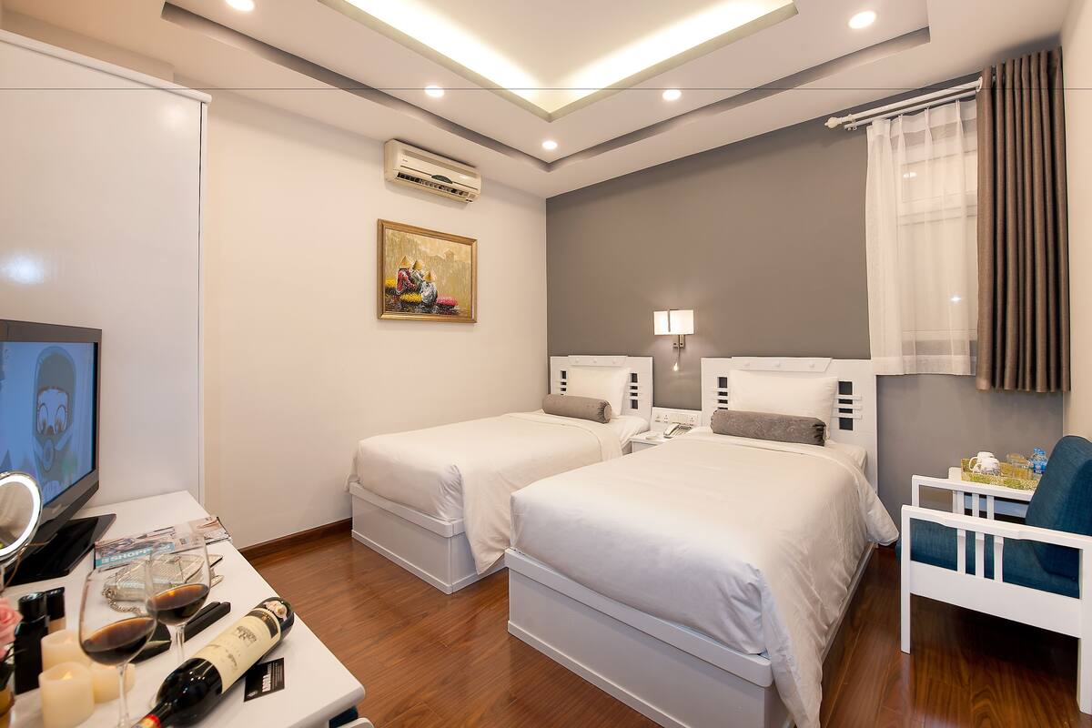 Une chambre d’hôtel avec deux lits simples, un bureau avec un ordinateur et un miroir, une télévision, et une petite table avec une bouteille de vin et des verres.