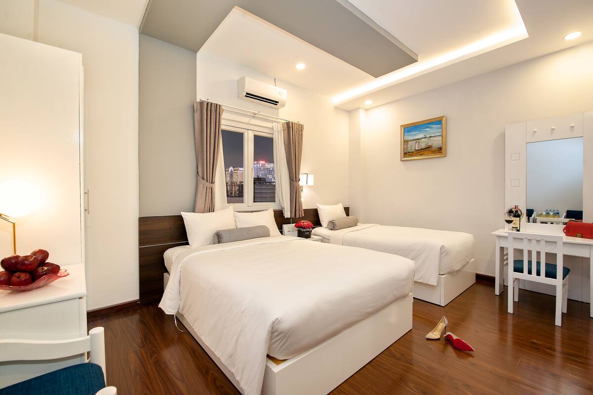 Une chambre d’hôtel avec deux lits, un bureau, une chaise, une fenêtre avec des rideaux, un climatiseur et un tableau au mur.