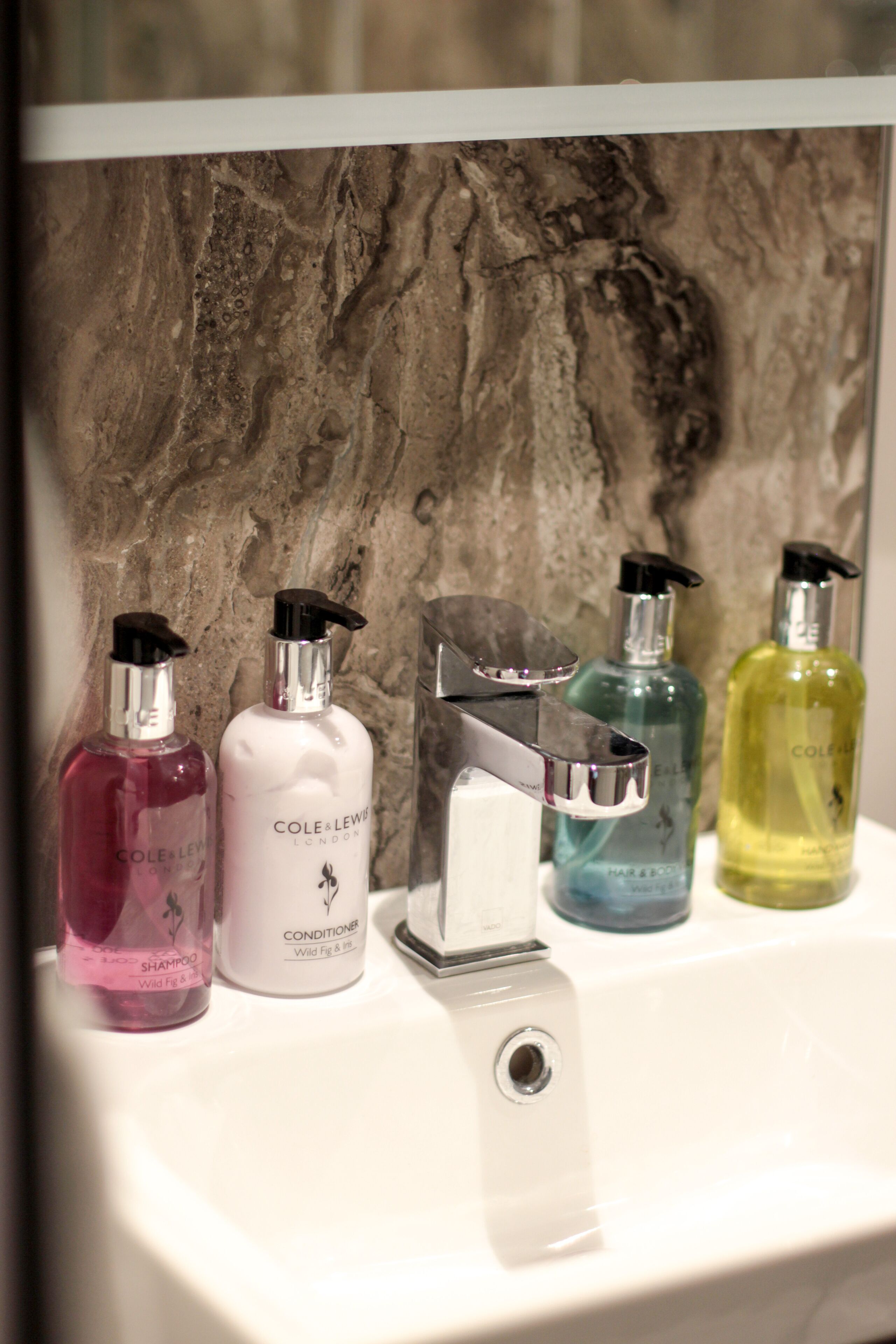 Une salle de bain avec un mur en marbre et un lavabo blanc. On y trouve cinq bouteilles de shampoing et d’après-shampoing posées sur le lavabo.