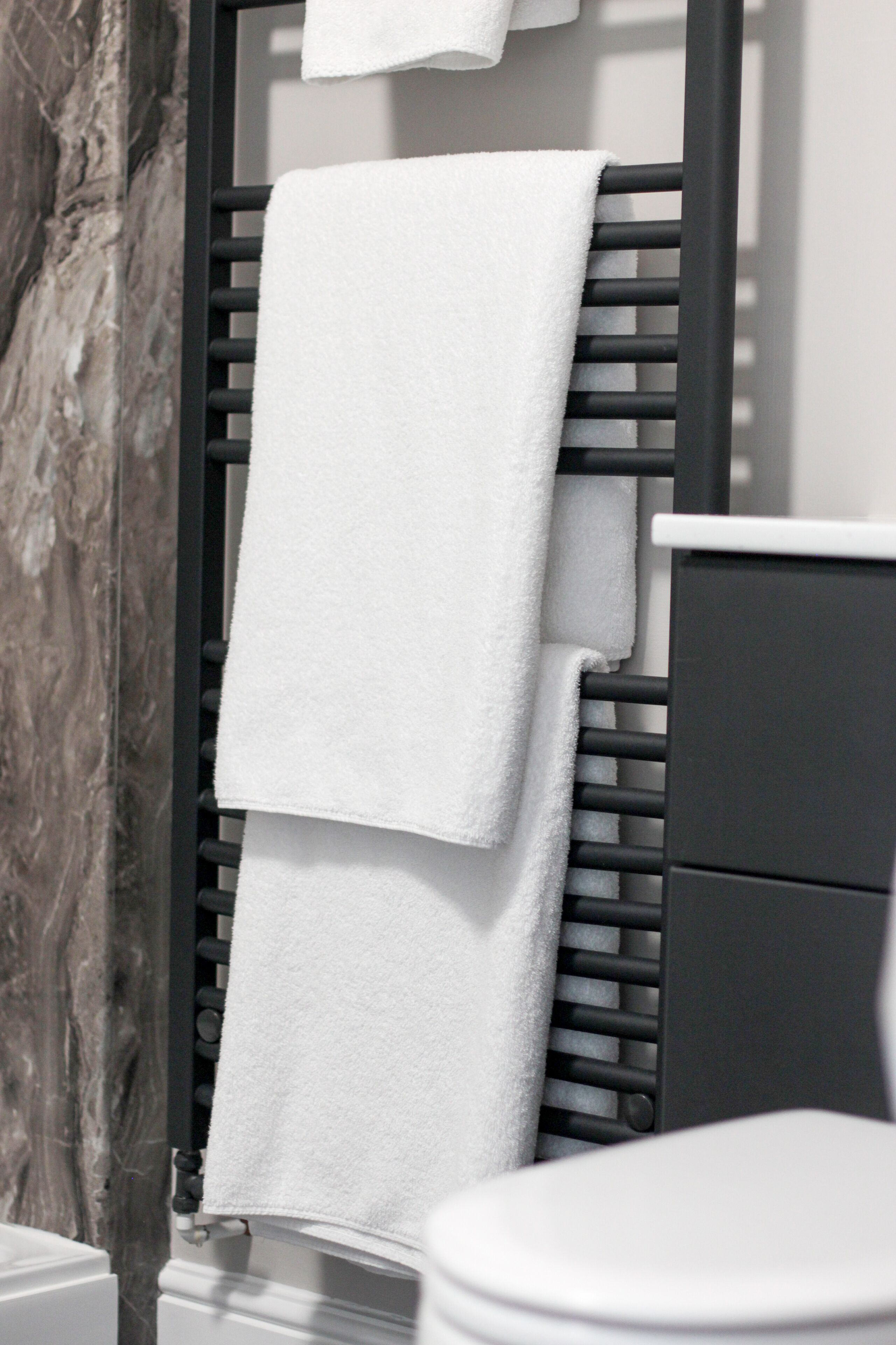 Un baño con un toallero negro que sostiene dos toallas blancas, un muro de mármol y un inodoro blanco.