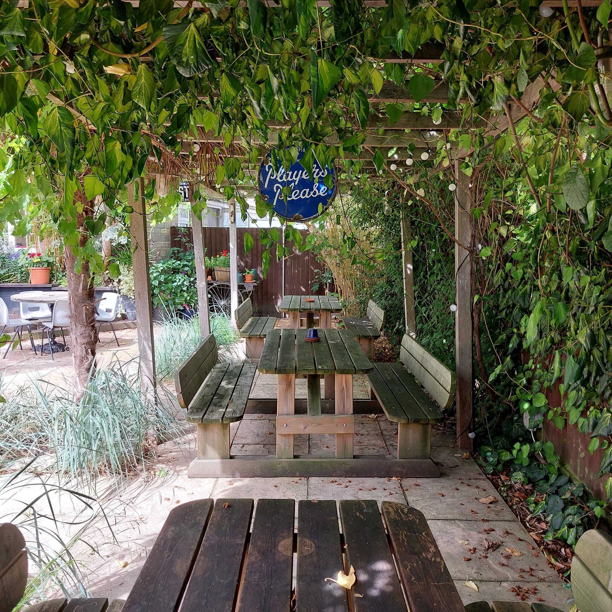 Una pérgola de madera con un letrero que dice "Play's Peace" y una mesa de picnic con bancos debajo.