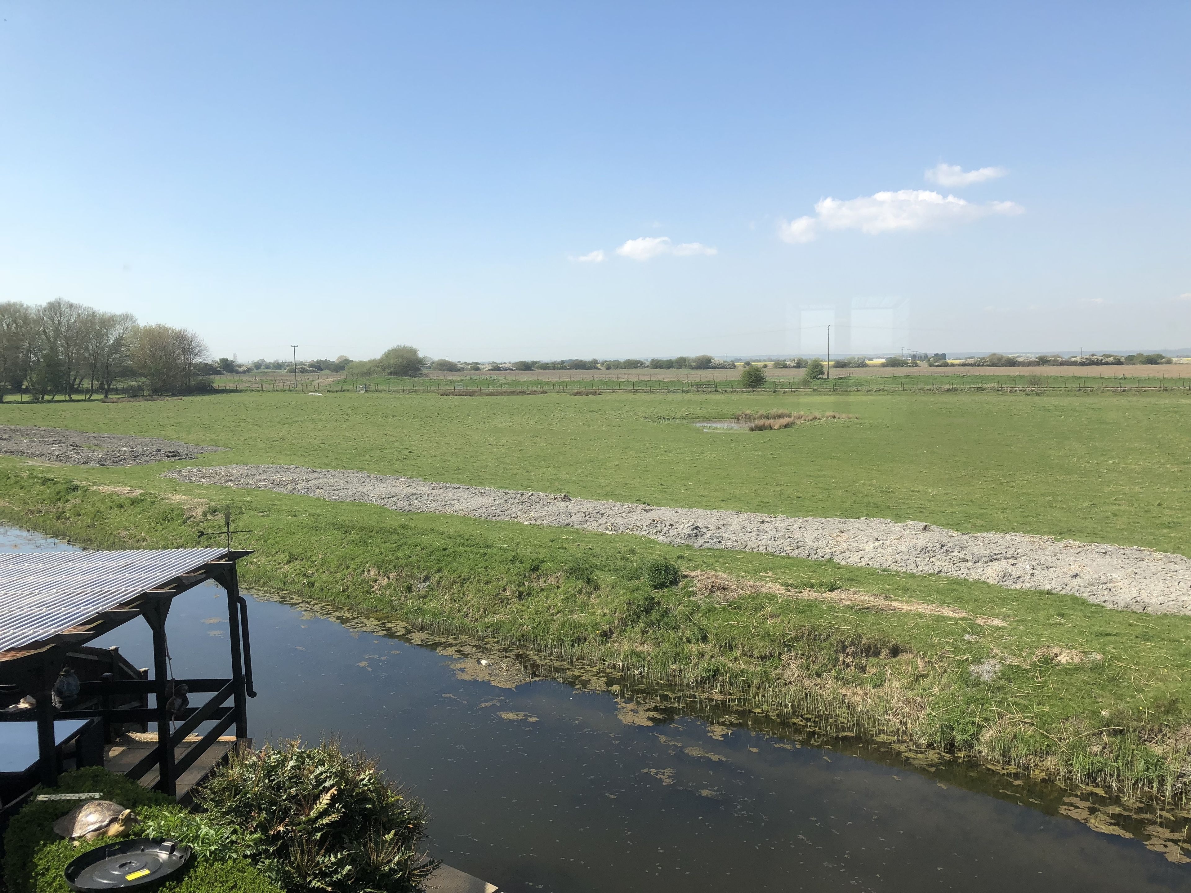A riverside view with a wooden dock and a small building, surrounded by open fields and a clear sky.