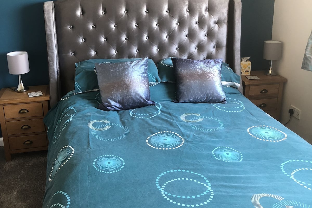 Ein Schlafzimmer mit einem gepolsterten Kopfteil, einer türkisfarbenen Bettdecke mit kreisförmigen Mustern, zwei Kissen, einem Nachttisch aus Holz mit einer Lampe und einem Beistelltisch mit einer Taschentuchbox.