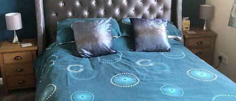 Un dormitorio con cabecera capitonada, colcha color turquesa con diseños circulares, dos almohadas, una mesita de noche de madera con una lámpara y una mesilla con una caja de pañuelos.