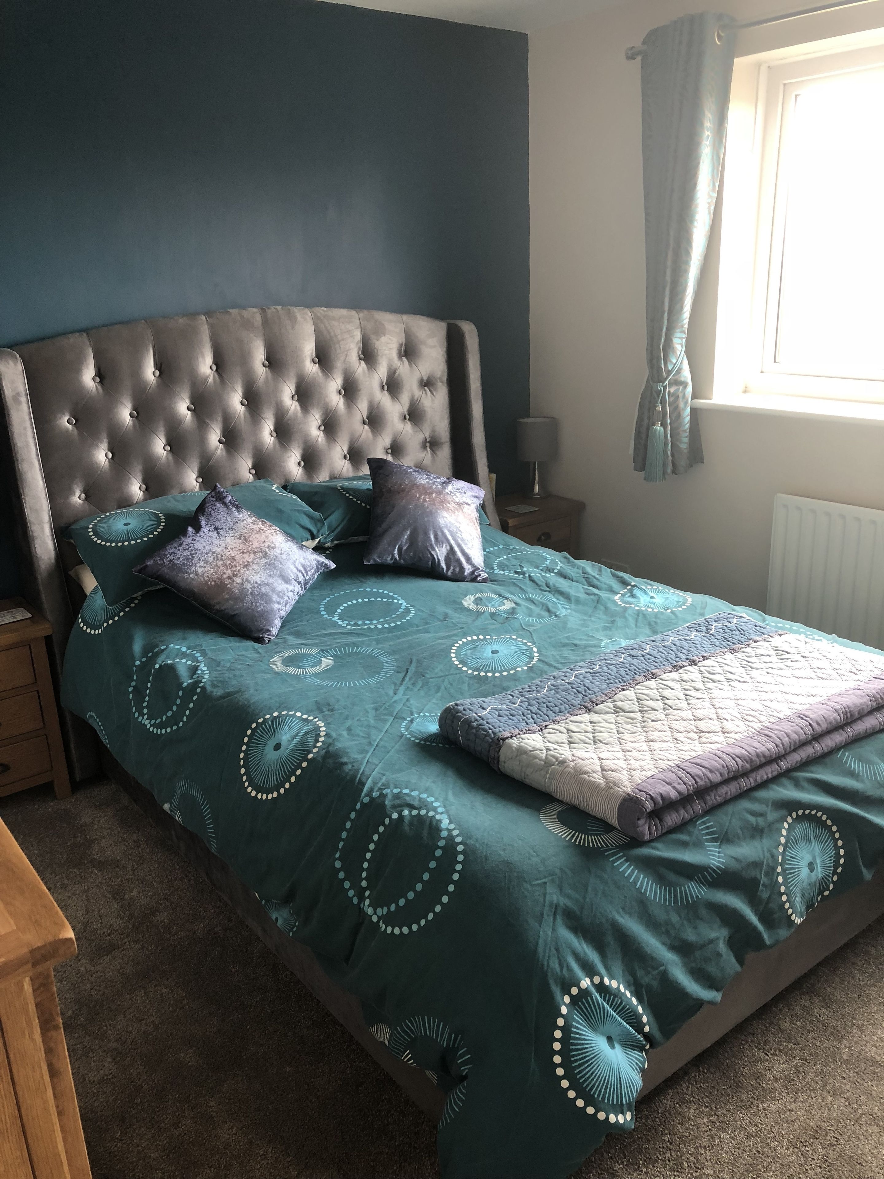 A bedroom with a tufted headboard, a bed with a patterned green and blue bedspread, and a window with light blue curtains.