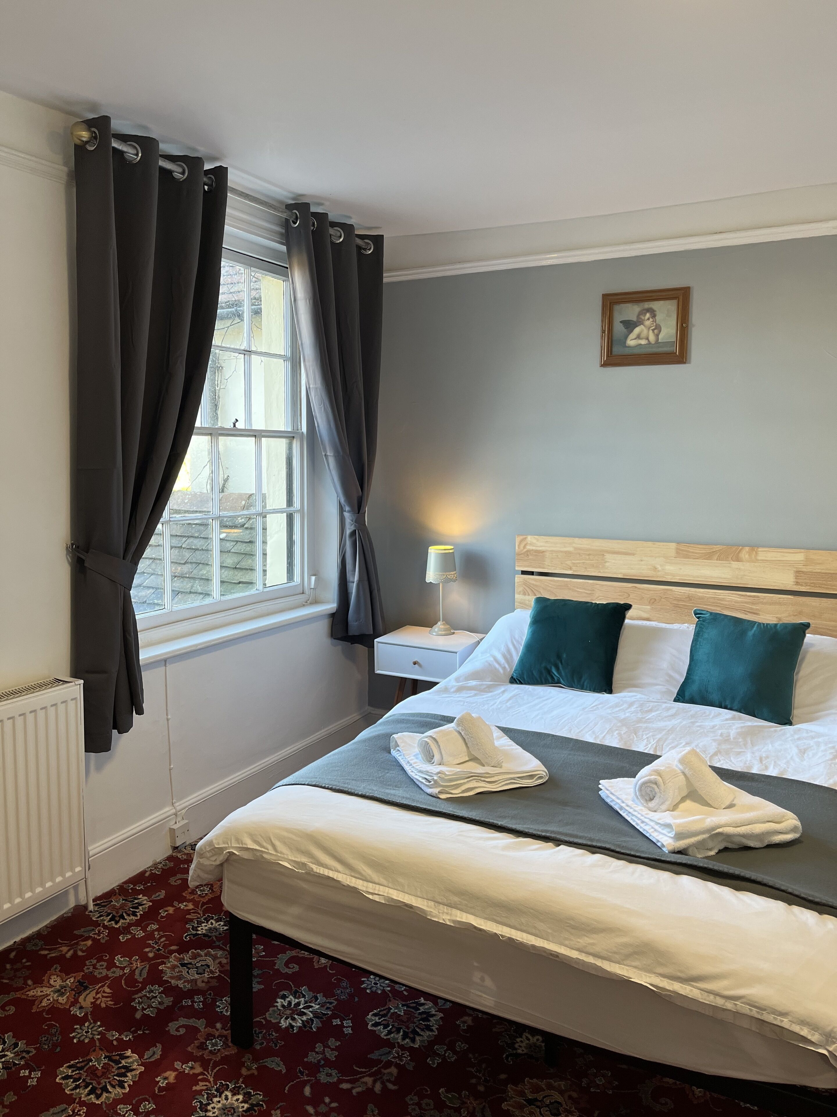 A neatly made bed with white linens and two teal pillows, a bedside table with a lamp, a radiator, and a window with dark curtains.