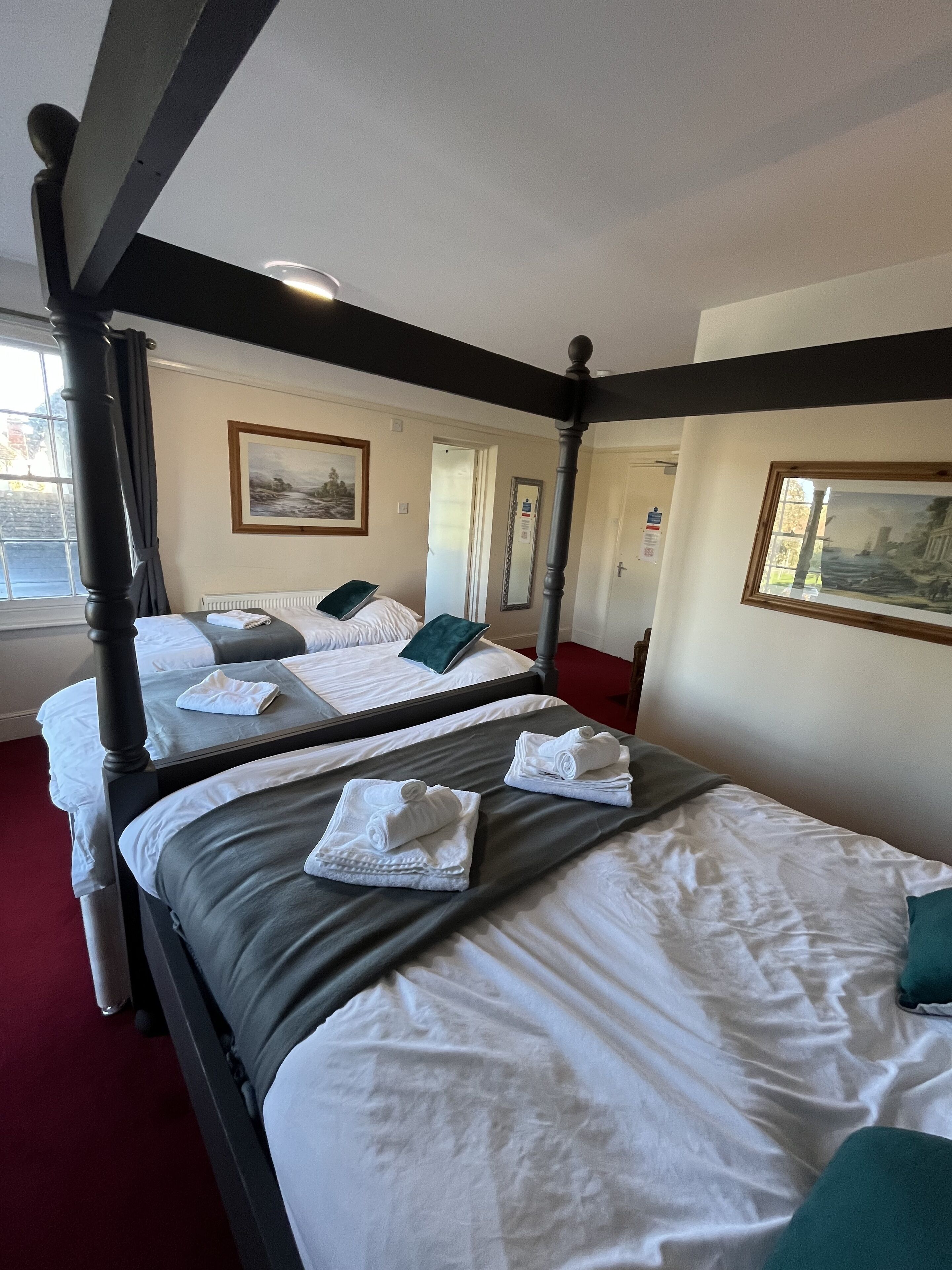Camera d'albergo con quattro letti singoli, ognuno con un asciugamano piegato e un cuscino verde. La stanza presenta una struttura letto a baldacchino in legno scuro e un'ampia finestra con vista sull'esterno.