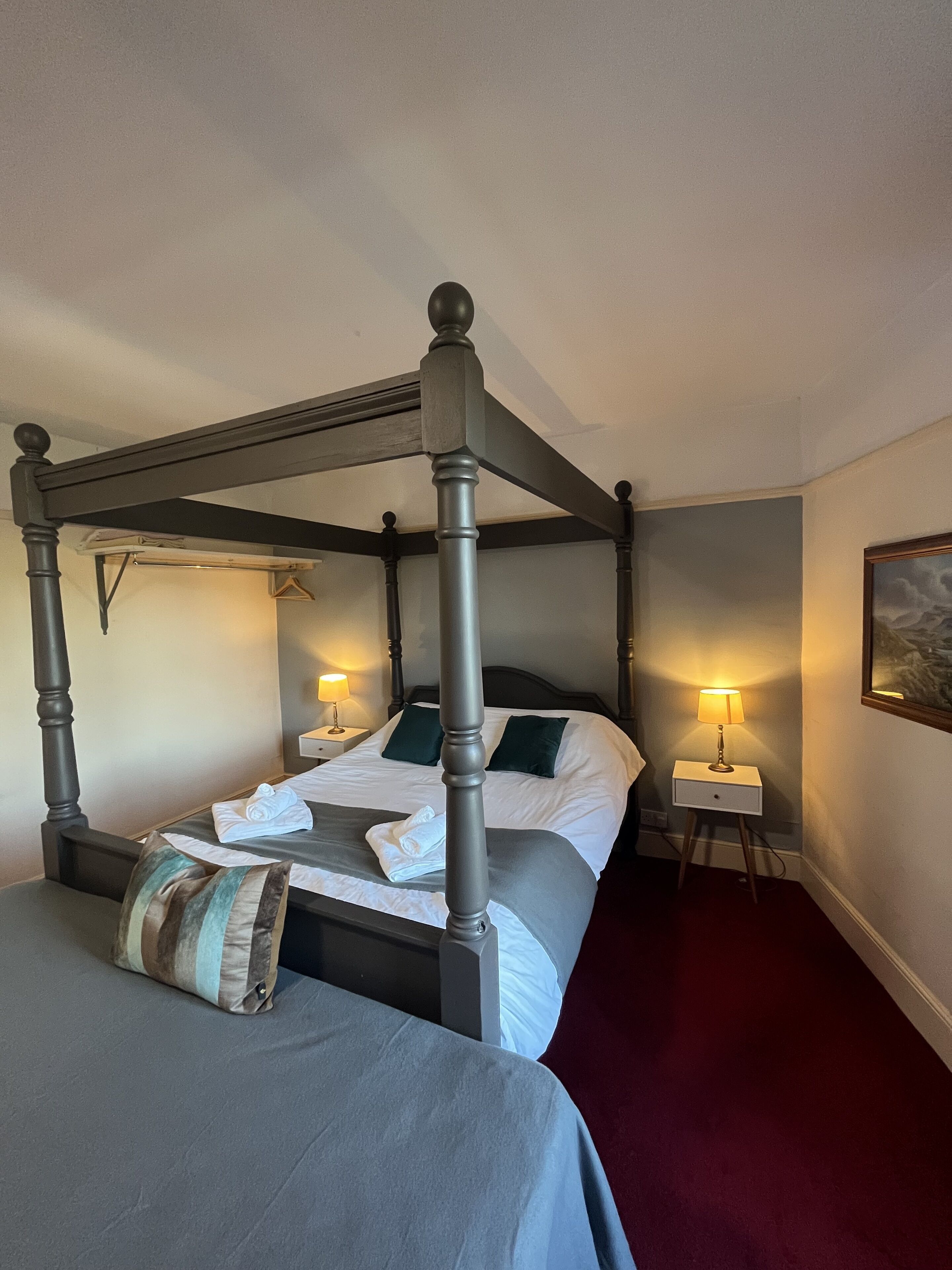 A four-poster bed with a grey headboard, white linens, and a striped pillow. There are two bedside tables with lamps, a red carpet, and a painting on the wall.