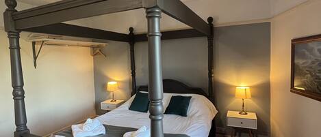 A four-poster bed with a grey headboard, white linens, and a striped pillow. There are two bedside tables with lamps, a red carpet, and a painting on the wall.