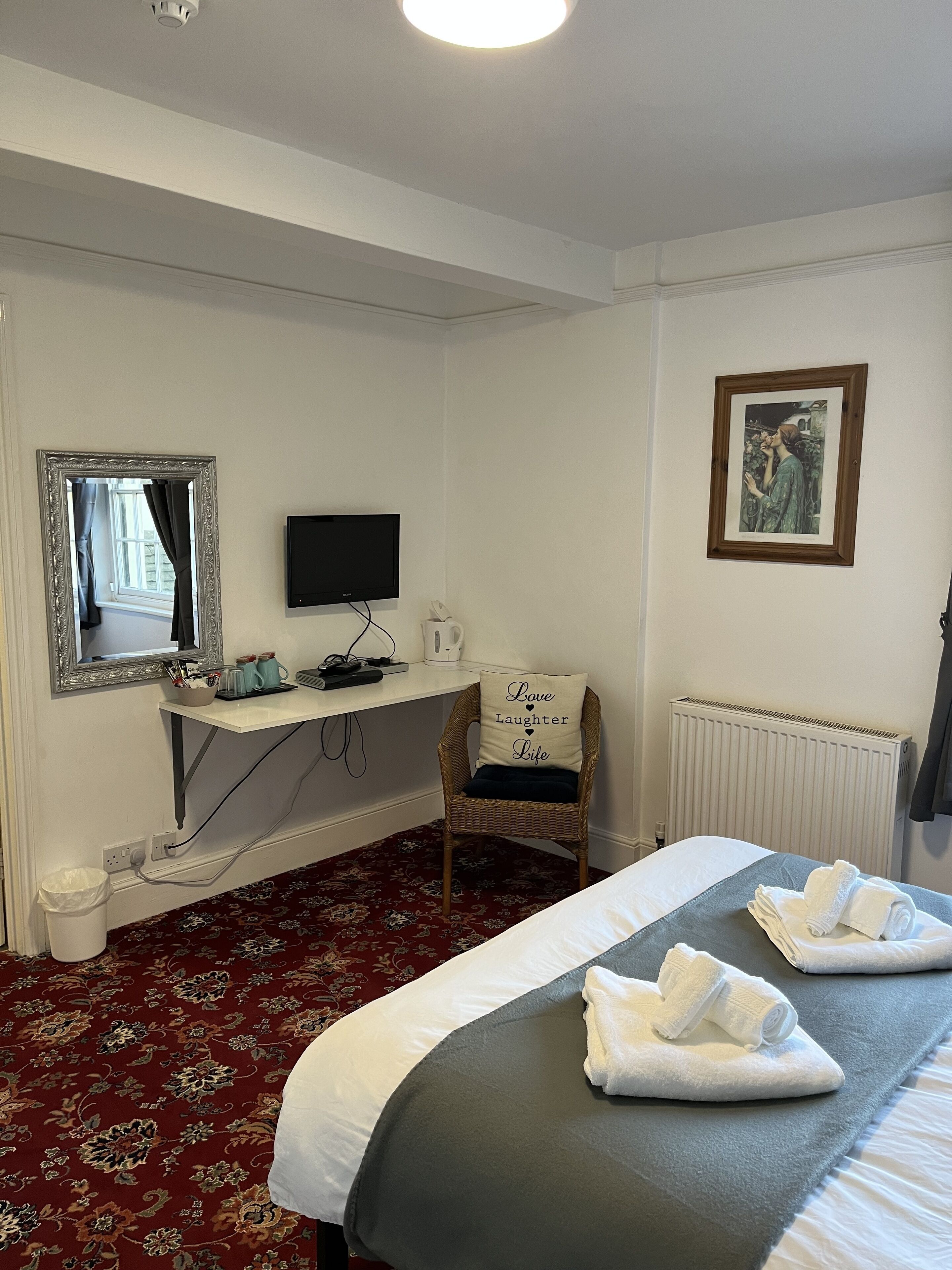 A hotel room with a bed, a chair with a pillow, a desk with a computer, a mirror, and a framed picture on the wall.
