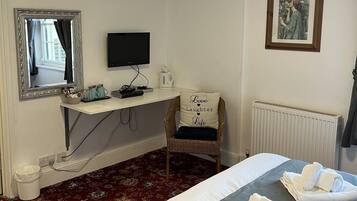 A hotel room with a bed, a chair with a pillow, a desk with a computer, a mirror, and a framed picture on the wall.