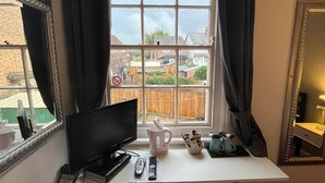 Quarto com escrivaninha, televisão, janela com cortinas e vista para uma área residencial.