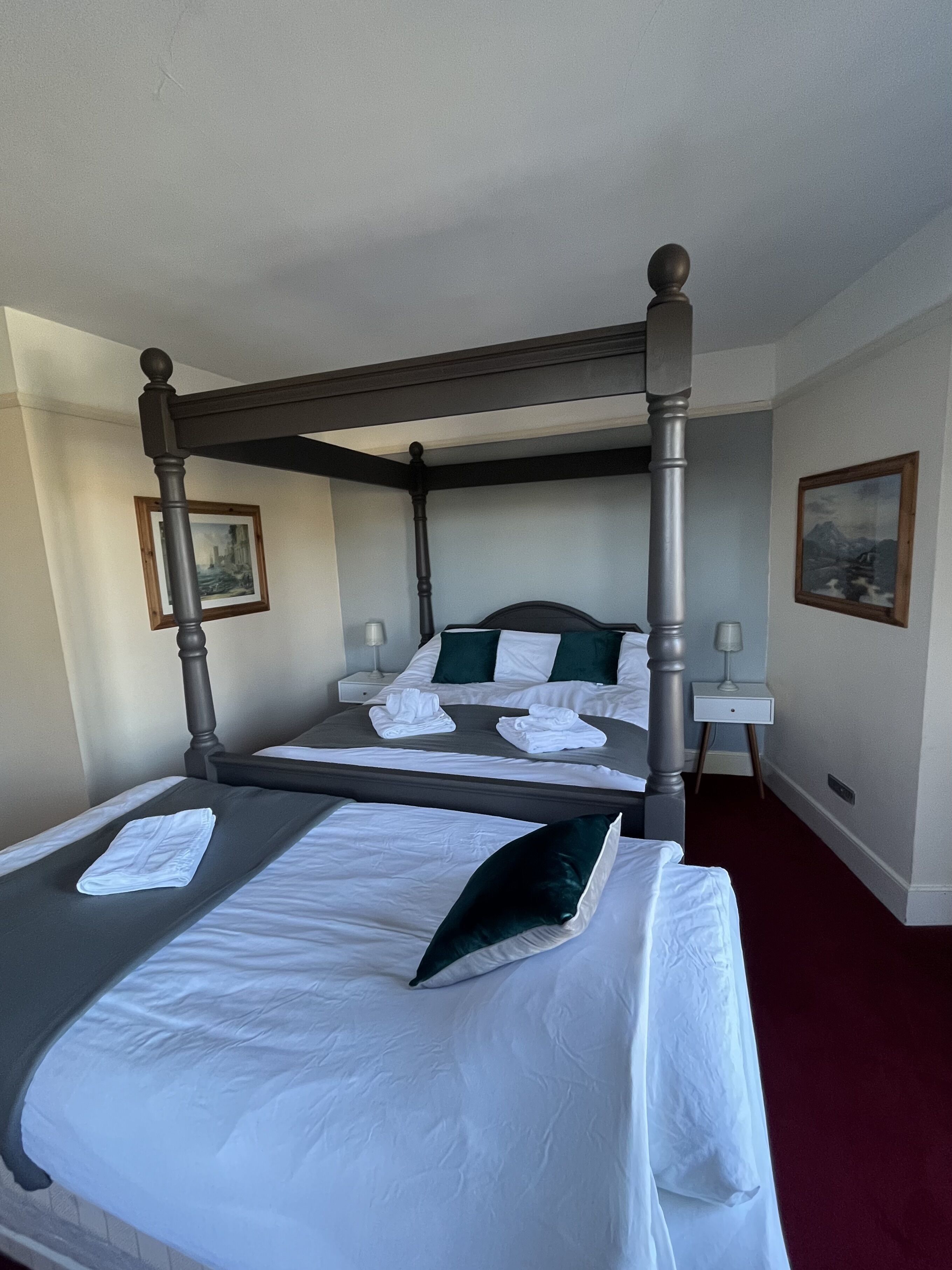 A four-poster bed with white linens and green pillows, a nightstand with a lamp, and framed pictures on the walls.