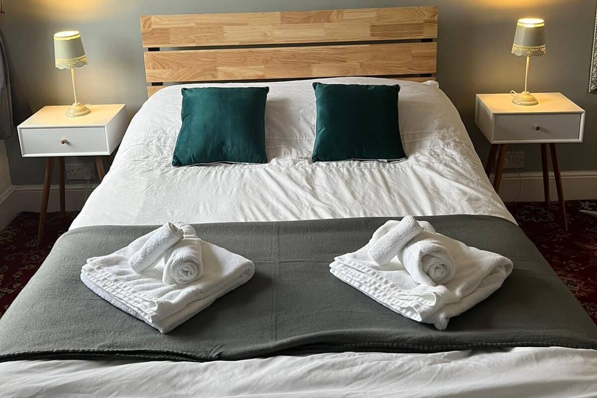 Una cama bien tendida con dos almohadas, dos mesitas de noche con lámparas y un cuadro en la pared.