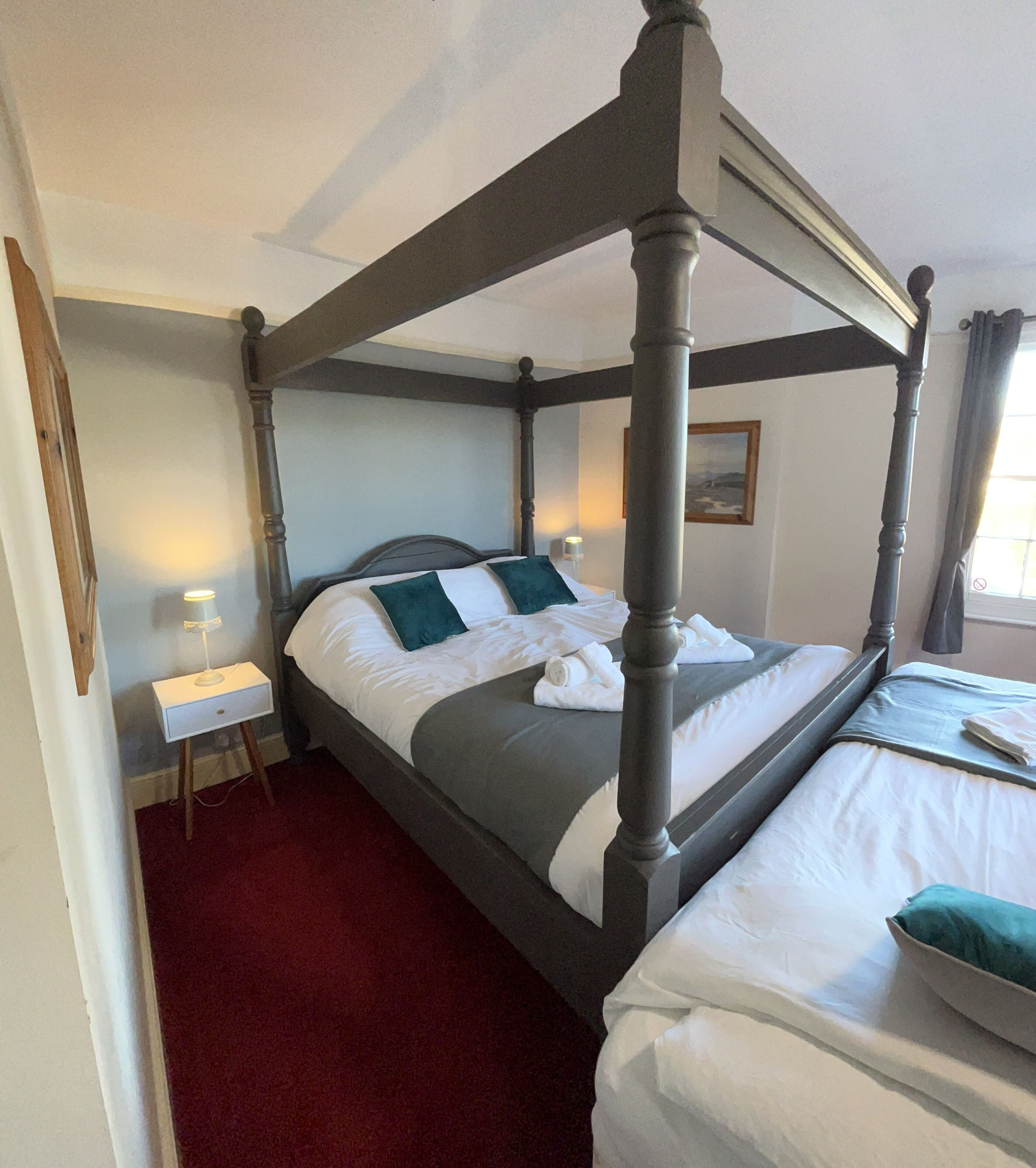 A four-poster bed with white linens and teal pillows, a bedside table with a lamp, and a red carpeted floor.