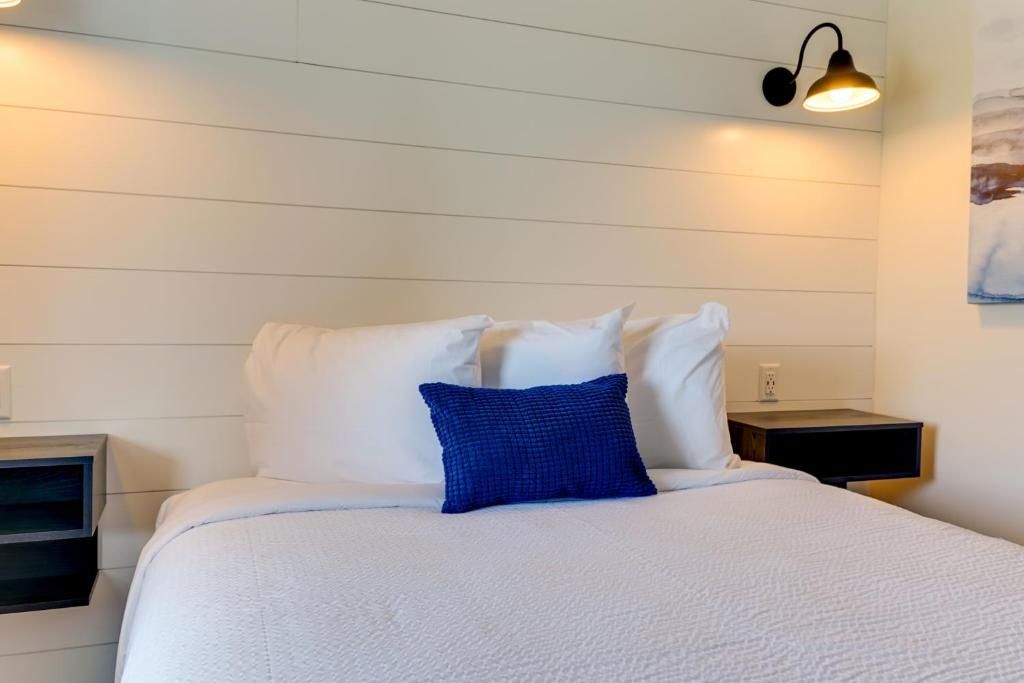 Una cama con ropa de cama blanca y una almohada decorativa azul, una mesita de noche con lámpara, y una lámpara de pared.