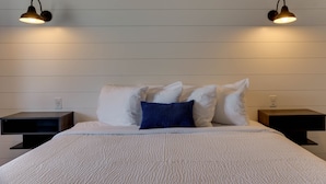A neatly made bed with white bedding and a blue pillow, flanked by two bedside tables with lamps, against a wall with horizontal paneling. - The Shore House (Ocean City)
