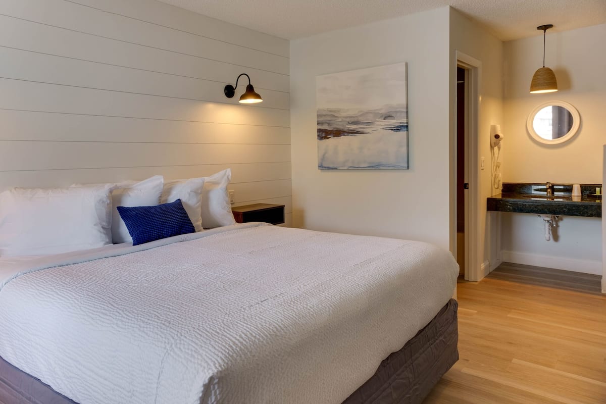 A bedroom with a large bed, a nightstand, a wall-mounted lamp, a painting, a round mirror, and a bathroom with a sink and countertop.