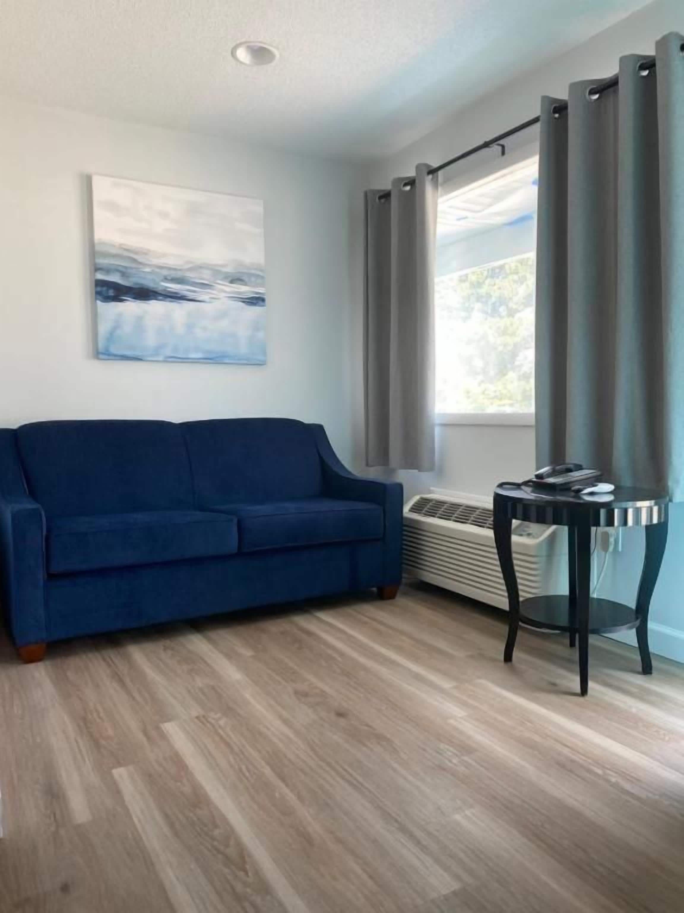 Une pièce avec un canapé bleu, une petite table, une fenêtre avec des rideaux et un climatiseur fixé au mur.