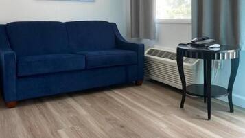 Une pièce avec un canapé bleu, une petite table, une fenêtre avec des rideaux et un climatiseur fixé au mur.